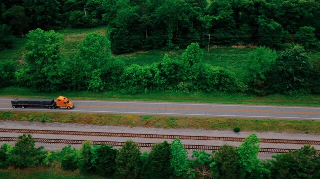 Flatbed semi hauling a full-length black tarped load down a rural two-lane highway, railroad tracks running parallel through green forest.
