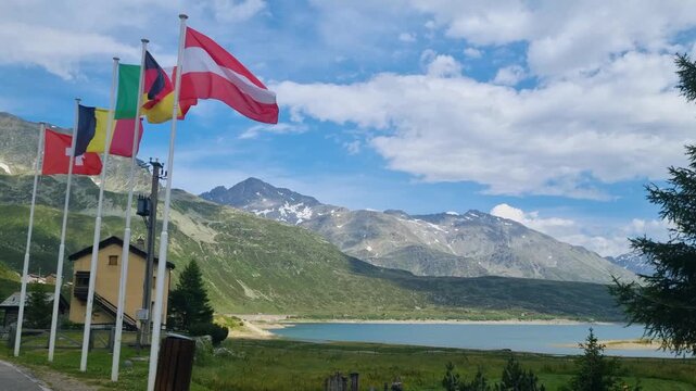 Flags of Austria, Germany, Italy, Belgium, and Switzerland sway above Lago di Montespluga on a cloudy day, symbolizing international travel in the Alps and the cross-border spirit of Europe's mountain