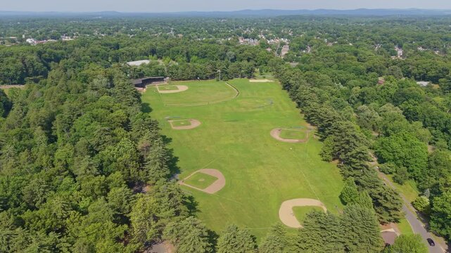 Drone orbit fly of several baseball diamonds sit in a wide green Athletic Field of Springfield bordered by dense trees and suburban neighborhoods, Massachusetts, US