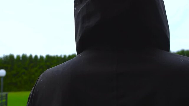 Slow motion view from behind of raindrops falling on the hood and shoulders of a person wearing a black waterproof jacket, with a blurry green garden and overcast sky in the background