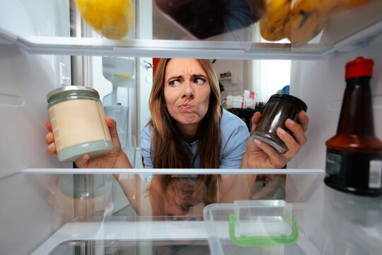 Confused Woman Looking Inside Refrigerator Choosing Between Two Jars. Undecided hungry female opens fridge door trying to select best food jar option 