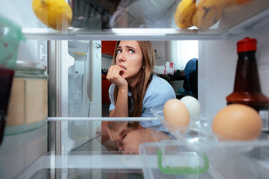 Anxious Hungry Woman Biting Nails Looking Inside Open Refrigerator. Stressed indecisive lady stares into kitchen fridge door while chewing fingernails checking food options