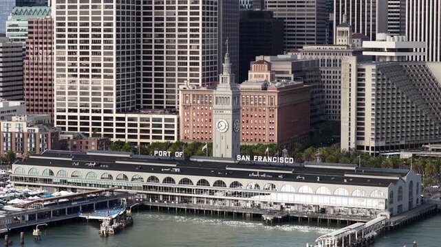 Aerial view showing the iconic Ferry Building Marketplace with its famous clock tower, set against the dense urban landscape of downtown San Francisco's modern skyscrapers