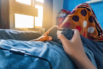 Close up view of gamer hands holding a black gamepad controller while relaxing on bed at home. Entertainment, digital technology, and cozy indoor lifestyle concept template. © Anzz Stock