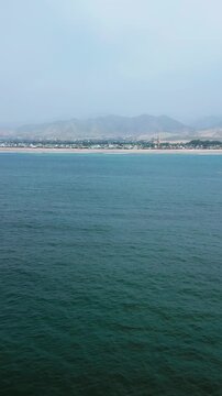 Vertical aerial drone footage flying forward over the Pacific Ocean toward the shoreline near Lima, Peru. Calm blue waters lead to a scenic sandy coast, creating a peaceful coastal landscape under bri