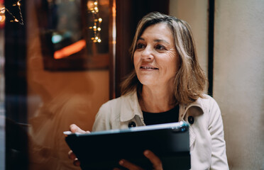 Middle aged woman holding digital tablet sitting in cafe. Thoughtful female entrepreneur planning business project using portable device gadget, financial investment, looking away, copy space © BullRun