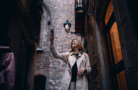 Mature woman traveler taking photo with smartphone in historic city alley. Happy tourist exploring old town architecture, digital nomadic lifestyle, wanderlust, authentic representation
