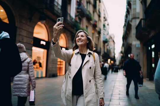 Happy mature woman holding smartphone for selfie in European city, middle aged traveler using mobile phone app for social media, authentic digital nomad lifestyle in urban setting.