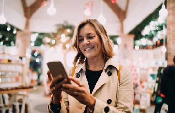 Cheerful adult woman checking smartphone notifications in festive market, smiling person using mobile app for online banking or shopping, authentic digital inclusion and modern lifestyle.