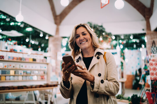 Smiling woman holding smartphone for online communication in festive interior, adult female using mobile device for remote work, authentic representation of digital nomad and technology literacy.