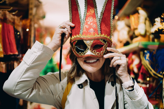 Smiling mature woman trying on ornate carnival mask in souvenir shop, happy middle aged female celebrating festival tradition, authentic portrait of joy and cultural discovery in retail store.