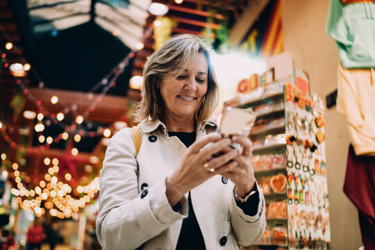Joyful mature woman looking at smartphone screen in city market, middle aged person enjoying mobile digital content, authentic portrait of tech savvy female using high speed internet app