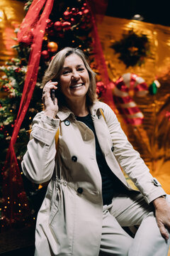 Joyful woman having mobile phone call during winter holidays, middle aged female professional using smartphone for communication, authentic portrait of digital lifestyle and seasonal celebration.