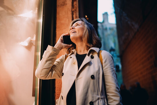 Happy mature woman calling smartphone night city, Caucasian lady looking up laughing during phone conversation, roaming connection, mobile communication outdoor, authentic people digital nomadism