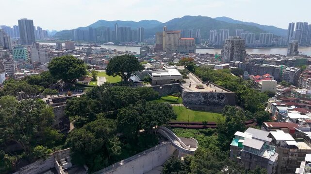 Macau Museum and historic Mount Fortress walls surrounded by trees tourists and dense city skyline beyond river - aerial parallax shot