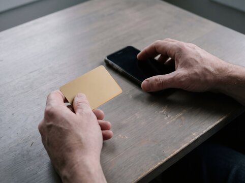 Male hands holding blank gold credit card and using smartphone on grey wooden table, digital transaction concept