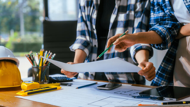 Two coworkers review building plans at a desk with a hard hat, tools, phone and tablet in a bright office, showing teamwork, planning and construction work.
