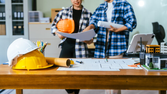 Two architects reviewing building plans in an office with hard hats, drawings, and a small house model on the desk