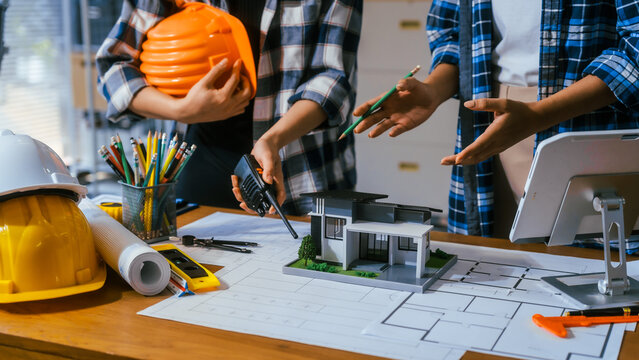 People discussing a house model with blueprints, tools, and safety helmets during an architecture project meeting