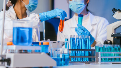 Fototapeta premium Masked laboratory technicians handling test tubes with blue liquid during scientific research in a modern lab