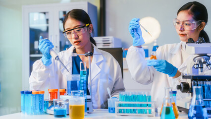 Fototapeta premium Two female scientists in lab coats and gloves use pipettes and microscope among test tubes and glassware in a modern laboratory.