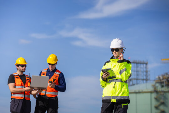 Female engineer with digital tablet and walkie talkie managing operations at lng plant, Professional industrial team in safety vests working with laptop and radio at liquefied natural gas facility