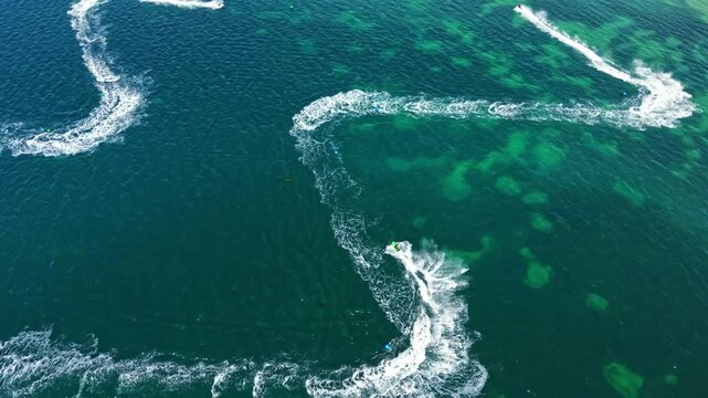 Racers ride jet skis on clear water while competing in a race. The scene shows waves being created as they turn and maneuver. It is daytime and the water is clear.