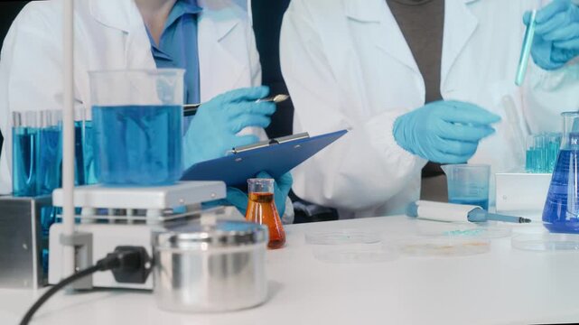 Two scientists in lab coats and blue gloves work with test tubes, beakers and a clipboard in a modern laboratory during an experiment.