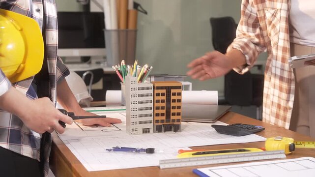 People planning a building project around a desk with blueprint, model buildings, tools and calculator, showing teamwork in architecture and construction