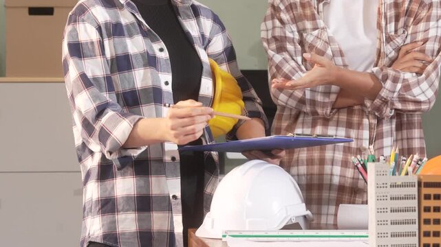 Two architects or engineers discussing plans with clipboard, hard hat, pencils and building model in an office setting