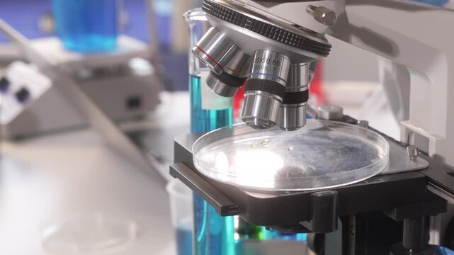 Close-up of a microscope over a petri dish in a laboratory, with blurred lab equipment and blue containers in the background.