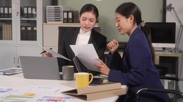 Two businesswomen reviewing documents during a meeting in a modern office with laptop, coffee mug, and paperwork