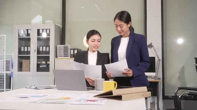 Two businesswomen reviewing documents together in a modern office with a laptop and desk papers, discussing work and planning a project
