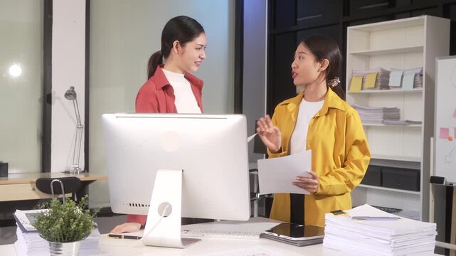 Two young women discussing work in a modern office, one holding papers beside a desktop computer and stacks of documents