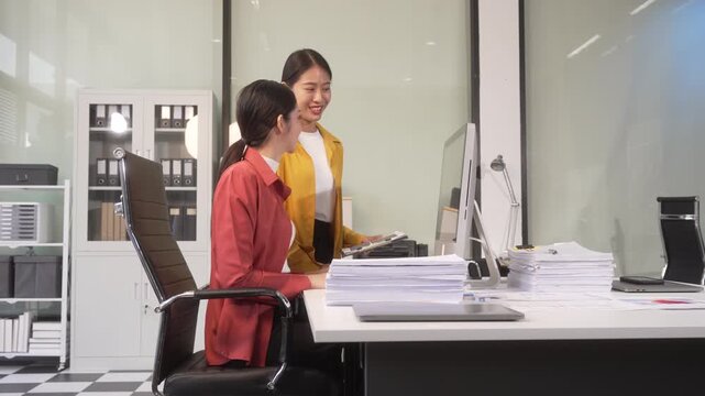 Two businesswomen in an office discussing work at a desk, with one pointing at a computer screen and stacks of paperwork nearby