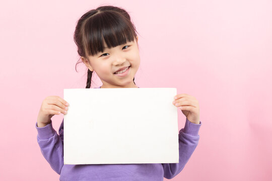 Happy young girl with braided hair holding a white empty card horizontally in front of a solid pink studio background.