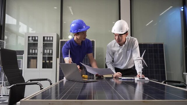 Two engineers in hard hats review plans and a laptop beside a solar panel model in a modern office meeting room