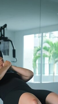 Vertical video. Young woman in black sportswear at the top position of a decline sit-up on a gym bench, facing the camera with a focused expression in a bright naturally lit gym space