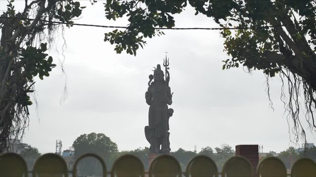 VADODARA, INDIA Static silhouette shot of the giant Lord Shiva statue at Sursagar Lake with foreground tree branches and passing vehicle traffic motion