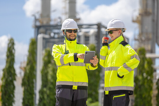Professional workers discussing operations with tablet and walkie talkie at petrochemical facility, smiling engineers in high visibility jackets reviewing data at oil and gas plant