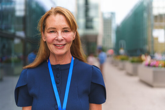 Senior businesswoman smiling, wearing a blue dress and lanyard outside an office building