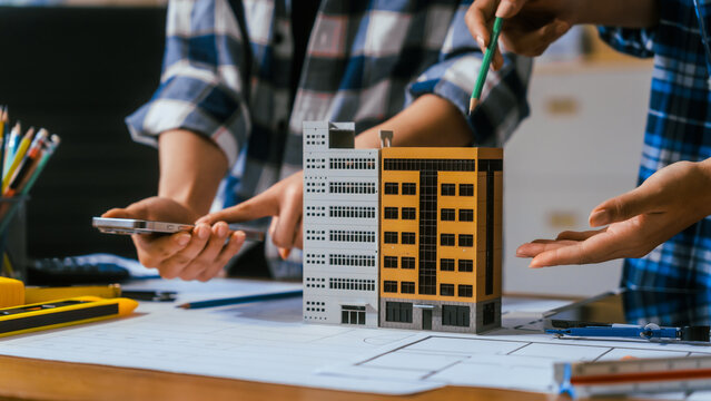 Architects review a small apartment building model over blueprints in an office, discussing design ideas with tools and a smartphone on the desk.