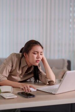 Tired young Asian businesswoman leaning on hand feeling exhausted while working on laptop at office desk