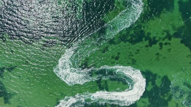 Racers ride jet skis on water creating waves and patterns. The sun brightens the scene as they speed around, enjoying the excitement of the race in a coastal area.