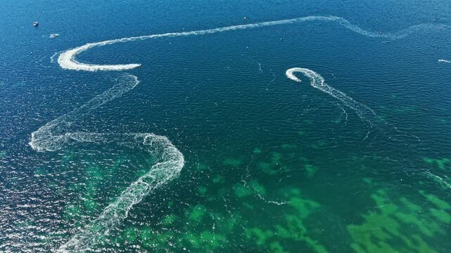 On a sunny day, jet ski racers zoom across the clear water, making sharp turns and creating waves. The water reflects the sunlight as the competitors race.