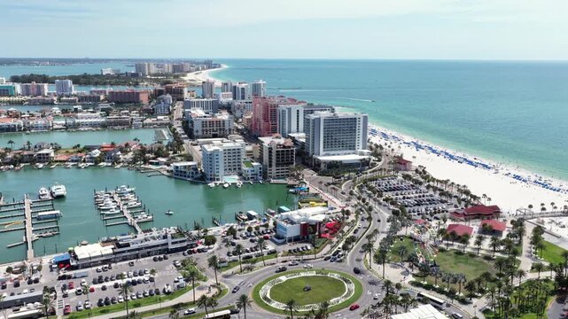 Palm-lined roundabout links marina district to white sand beach in Clearwater Beach, Florida, high-rise hotels overlook rows of blue beach umbrellas near turquoise water. Aerial drone wide shot