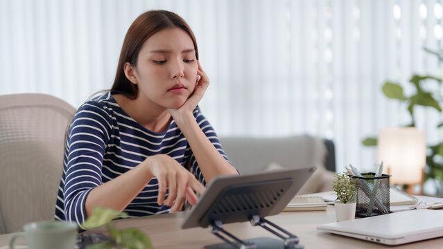 Young Asian woman looking bored and unmotivated using tablet at home workspace with copy space