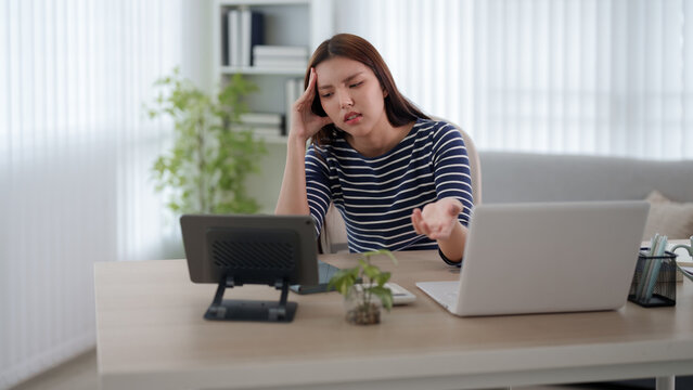 Stressed young Asian woman experiencing work burnout while multitasking with laptop and tablet at home office desk