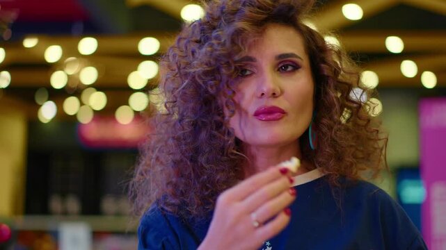 Woman with curly hair holds popcorn in a bright indoor space. She gazes directly at the camera with a curious expression. Soft, warm bokeh lighting creates a cozy atmosphere