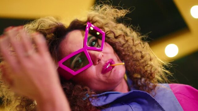 Woman adjusts her bold pink glasses playfully. Voluminous curls frame her smiling face. Yellow straw rests in her mouth. Warm, glowing string lights create a festive background